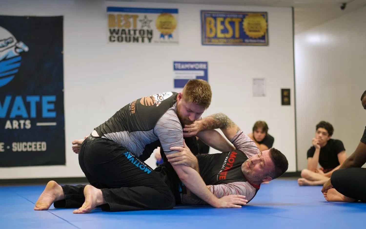 Two adults grappling No-Gi in rashguards on the blue mats while students watch